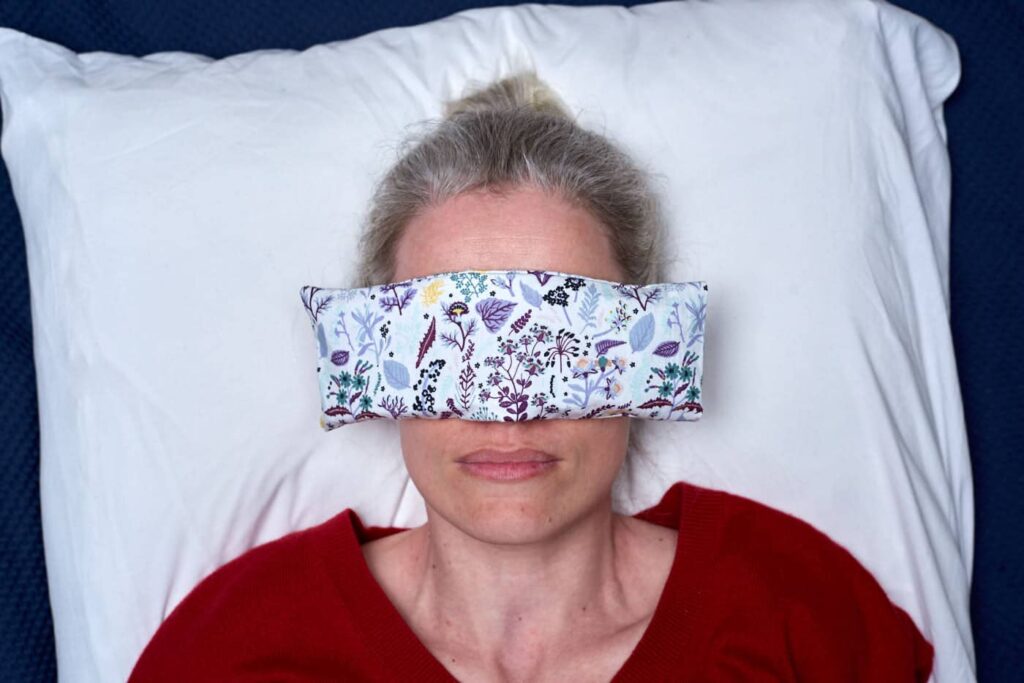 head shot of woman laying on white pillow, an eye pillow covers her eyes